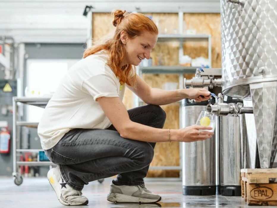 Worker tapping water kefir from a stainless-steel fermenter into a glass at a small brewery.